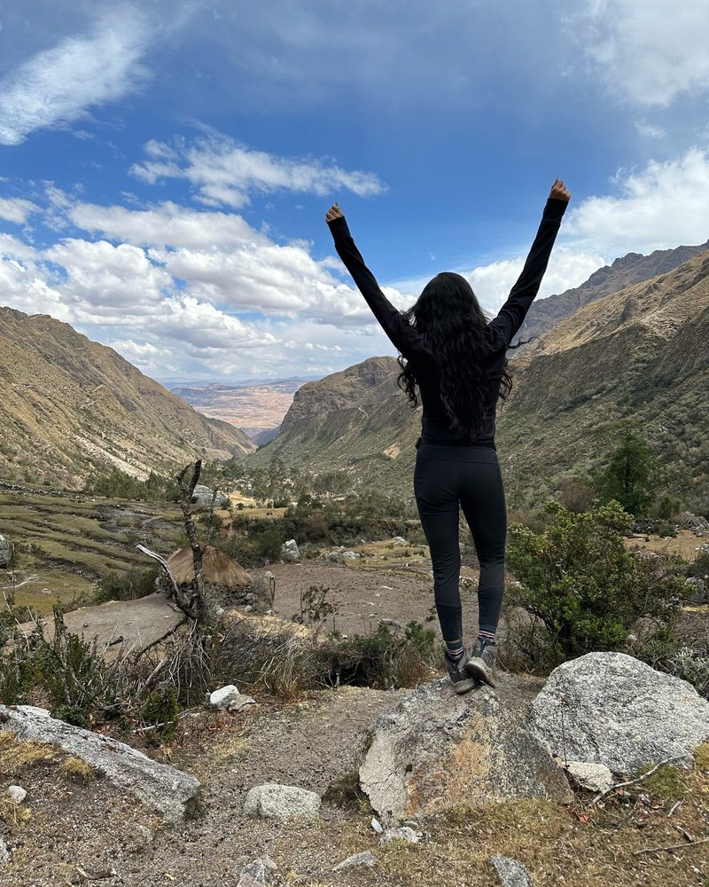 A person standing on a boulder with both arms raised, looking down a green valley toward distant plains