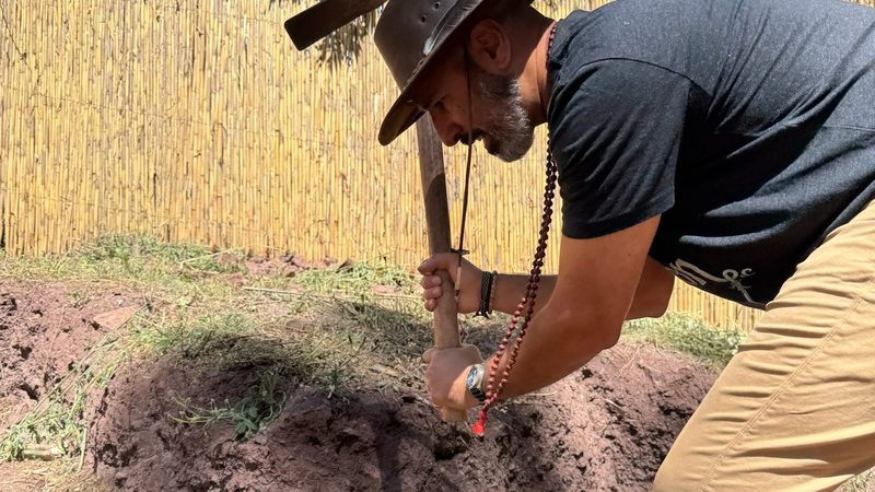 A person in a wide brimmed hat driving a pickaxe into a bank of red earth, reed fencing behind