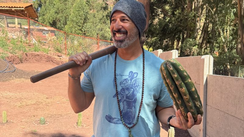 A person with a pickaxe over one shoulder carrying a Huachuma cutting under the other arm across the garden