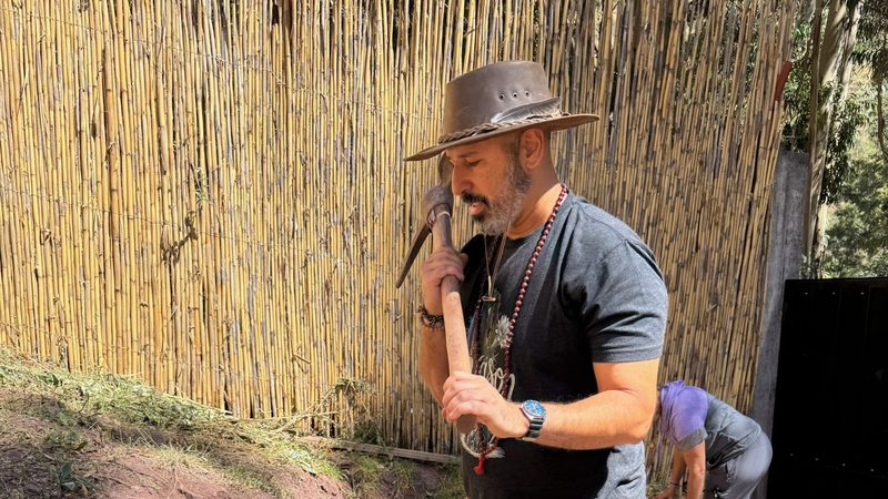 A person in a wide brimmed hat carrying a pickaxe against one shoulder along a reed fence in the garden, someone bent at work behind them