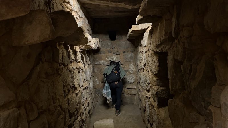 A narrow stone passage running under the temple at Chavín de Huántar, roofed with heavy slabs and lit by one small lamp, a person standing at the far end