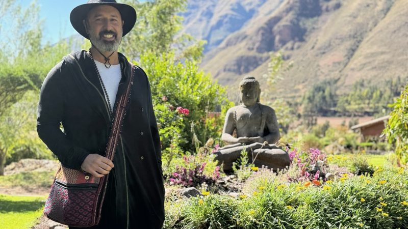 A person standing on a lawn beside a flower bed, a stone seated figure and a steep mountain ridge behind
