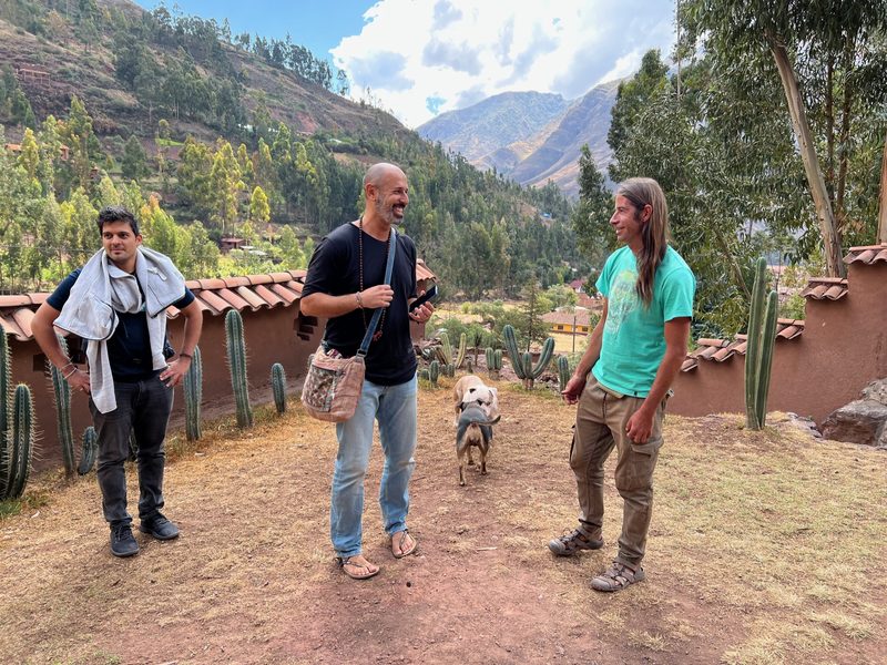 Three people standing talking on a dirt yard beside an adobe wall topped with cactus, a dog walking between them