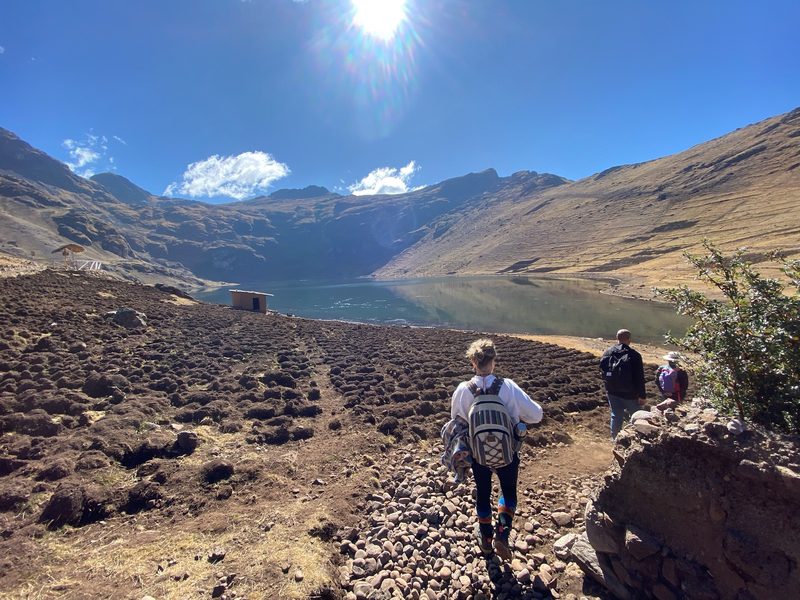 Three people walking down a stony path toward a lake, a small adobe hut standing at the water's edge
