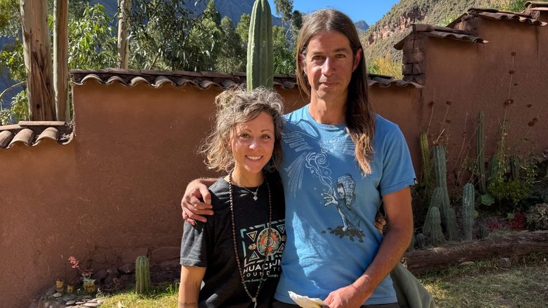 Two people standing together in a walled courtyard, a Huachuma column and the valley slope behind them
